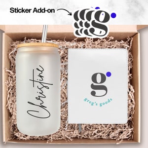 Pode incluir: Uma caixa de presente branca cheia de aparas de papel castanho. Dentro da caixa há um copo transparente com tampa de bambu e um canudo, personalizado com o nome "Christine". A caixa também contém um caderno branco com um logotipo "g" cinzento e o texto "greg's goods" em turquesa.