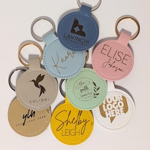 May include: A collection of personalized round keychains in various colors, including blue, pink, yellow, and beige. Each keychain features custom text or logos, such as names, company logos, and the text "YOUR LOGO HERE".