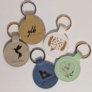 Puede incluir: Una colecci&oacute;n de cinco llaveros redondos de cuero en varios colores, cada uno con un anillo met&aacute;lico. Los llaveros presentan logotipos y texto, incluyendo "ylh YOUR LOGO HERE", un colibr&iacute;, "YOUR LOGO HERE", "LAVINCHI TECHNOLOGY" y "The Smith Family".