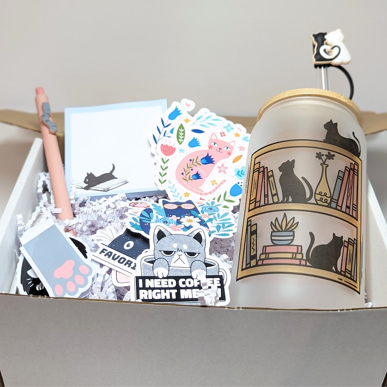 May include: A white gift box filled with various cat-themed items, including stickers, a pen, and a frosted glass tumbler with a cat design. The tumbler has a black cat silhouette and the text "I need coffee right meow".
