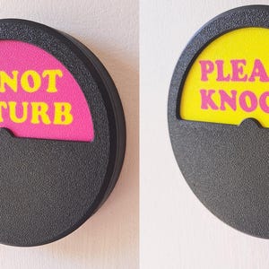 May include: Two round, black, plastic signs with a pink knob on the side. The signs have a yellow and pink semi-circle with text. The left sign says "DO NOT DISTURB" in yellow letters on a pink background. The right sign says "PLEASE KNOCK!" in pink letters on a yellow background.