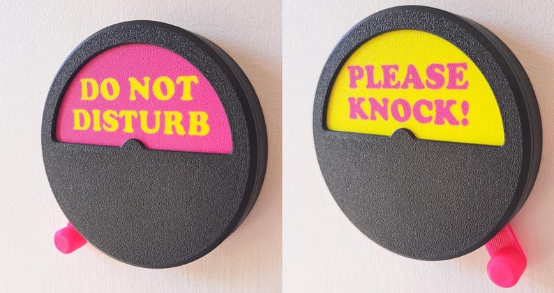 May include: Two round, black, plastic signs with a pink knob on the side. The signs have a yellow and pink semi-circle with text. The left sign says "DO NOT DISTURB" in yellow letters on a pink background. The right sign says "PLEASE KNOCK!" in pink letters on a yellow background.