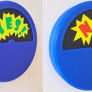 May include: A blue, round, wall-mounted, spinning decision maker with a black background and a yellow starburst. The starburst has the word "YES" in green letters on the left and the word "NO" in red letters on the right.