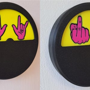 May include: Two black and yellow round wall-mounted signs with a pink hand gesture on each. The left sign has a pink hand making the rock and roll hand gesture. The right sign has a pink hand making the middle finger gesture. Both signs have a pink lever on the side.