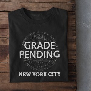 Könnte beinhalten: Schwarzes T-Shirt mit einer weißen Grafik des New Yorker Stadtsiegels mit dem Text "GRADE PENDING" darüber und "NEW YORK CITY" darunter.