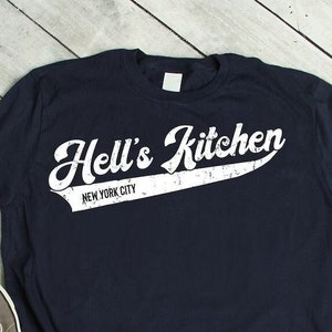 May include: A navy blue t-shirt with a white graphic that says "Hell's Kitchen New York City" in a vintage baseball font.