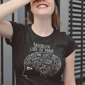 Op de afbeelding: Een zwart T-shirt met een witte afbeelding van een brein dat is verdeeld in secties die verschillende buurten in Brooklyn, New York, voorstellen. De tekst "Brooklyn State of Mind" en "Park Slope" is zichtbaar op de afbeelding.
