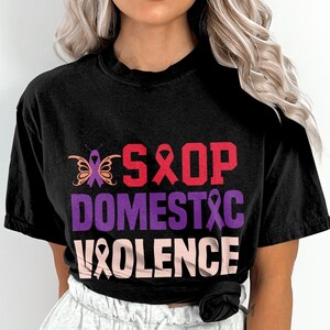 Könnte beinhalten: Schwarzes T-Shirt mit dem Text "STOP DOMESTIC VIOLENCE" in lila, rosa und roten Buchstaben. Ein Schmetterling mit einem lila Band befindet sich über dem Text.