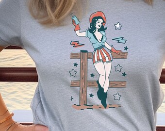 May include: Gray t-shirt with a vintage style illustration of a woman in a red hat and a red, white, and blue skirt standing on a wooden fence. The illustration is surrounded by stars and lightning bolts.