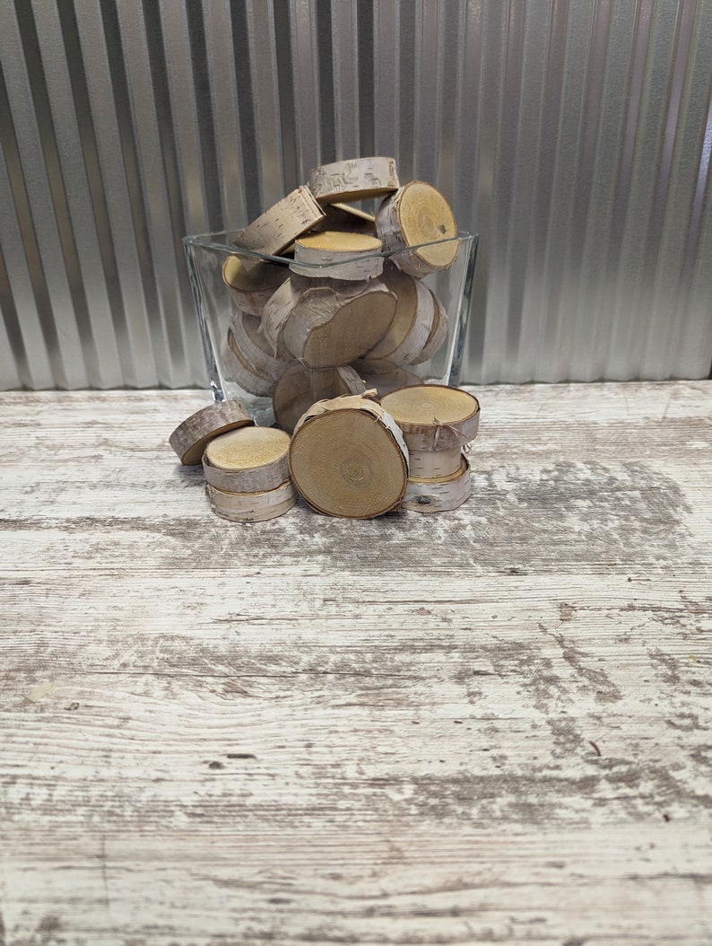 May include: A glass vase filled with natural birch wood slices. The vase is sitting on a whitewashed wooden surface.