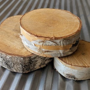 May include: Three natural wood coasters made from birch tree slices. The coasters have a light brown, circular surface with a textured, white and tan bark edge. The coasters are stacked on top of each other.