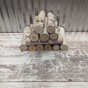 May include: A stack of small, white birch logs arranged in a pyramid shape. The logs are on a whitewashed wooden surface.