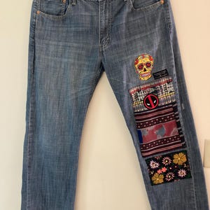May include: Light blue denim jeans with decorative patches. Patches include a colorful sugar skull, a Deadpool logo, and floral and patterned fabric designs. The jeans have a classic five-pocket design.