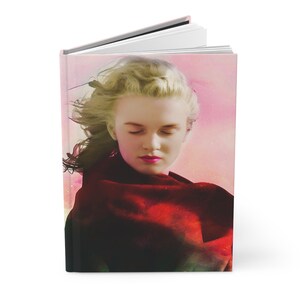 May include: Hardcover journal featuring a portrait of a woman with closed eyes and flowing blonde hair. The cover has a pink and red color scheme, with a red shawl. The journal is open, revealing white pages.