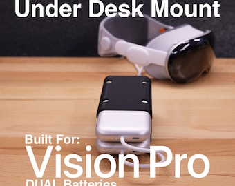 May include: Under Desk Mount for Vision Pro with dual batteries. The mount is black and has a white cable attached to it. The Vision Pro headset is white and gray and is sitting on a wooden surface.