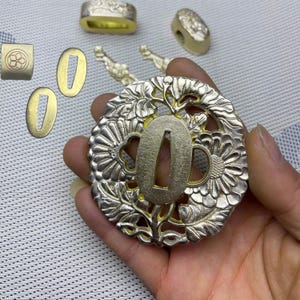 May include: A collection of Japanese sword fittings, including a tsuba with floral and leaf designs, and other decorative metal pieces. The tsuba is silver-colored with a gold-colored center.