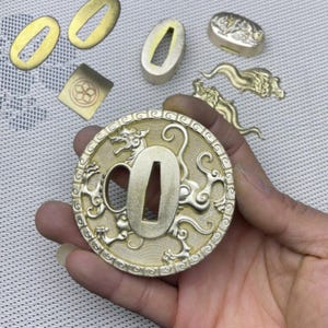 Peut inclure: Une garde d'épée en métal doré avec un motif détaillé d'un dragon avec des nuages tourbillonnants. La garde est circulaire et possède une bordure décorative.