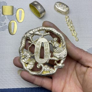 May include: A gold-coloured metal Japanese sword hilt, or tsuba, with a detailed design of two dragons and clouds. The tsuba is a circular shape with a rectangular opening in the centre. The tsuba is surrounded by other gold-coloured metal pieces, including two oval-shaped pieces, a rectangular piece, and a long, thin piece with a decorative design.