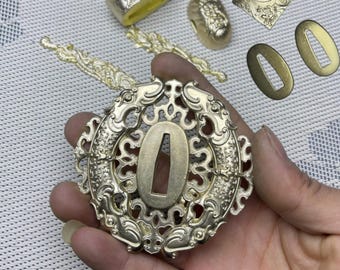 May include: A gold-colored metal sword guard with a detailed design of two fish and intricate floral patterns. The guard has a circular shape and a central opening for the sword blade.