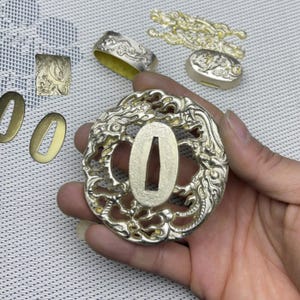 May include: A gold-coloured metal tsuba, a decorative sword guard, with a detailed design of a dragon. The tsuba is round and has a central oval opening for the sword blade.
