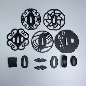 May include: A set of ten black metal sword fittings, known as tsuba, with various intricate designs. The tsuba are used to protect the hand from the blade.