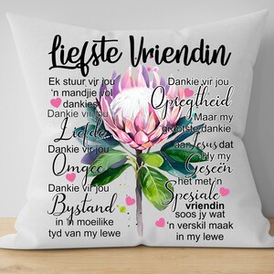Puede incluir: Una almohada blanca con un diseño de flor de protea rosa y morada y texto en holandés que dice "liefste vriendin" e incluye las palabras "dankie vir jou" y "omgee".