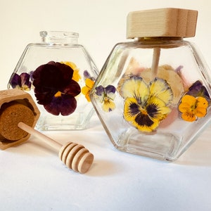 May include: Two clear glass hexagonal honey jars with wooden lids and a wooden honey dipper. The jars are filled with honey and decorated with pressed pansies.