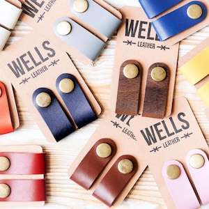 May include: A selection of leather tabs in various colours, each with a brass rivet, presented on cards branded with "WELLS LEATHER". The tabs are in shades such as white, grey, blue, yellow, brown, red, pink, and green.