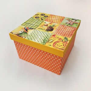 May include: A square-shaped box with a patchwork design featuring a scarecrow, a pumpkin, a hedgehog, and sunflowers. The box is orange with white polka dots and has a yellow lid.
