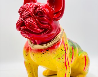 May include: A red and yellow ceramic figurine of a French bulldog. The bulldog is sitting with its head tilted to the side. The figurine is painted with a yellow base and red accents, with pink and green splatters of paint.