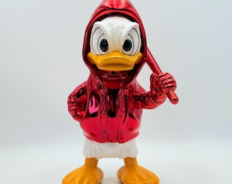May include: A red metallic Donald Duck figurine holding a baseball bat. The figurine is wearing a red hooded jacket and has a white chest, yellow feet, and a yellow beak. The figurine is standing on a white surface.