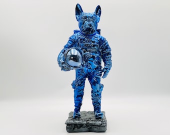 Könnte beinhalten: Eine blaue, metallische Figur eines französischen Bulldoggen, der als Astronaut verkleidet ist und einen Helm hält. Die Figur steht auf einem schwarzen Sockel.