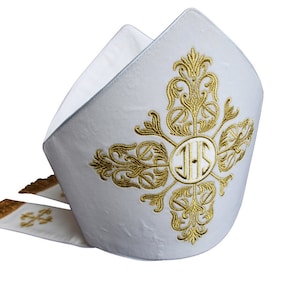 Puede incluir: Mitra blanca con bordado dorado. El bordado presenta una cruz con un círculo en el centro que contiene las letras "IHS".