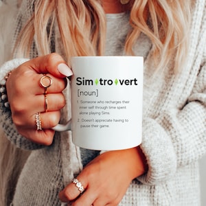 May include: A white ceramic mug featuring the black text Sim-tro-vert [noun] with green diamond icons. The definition reads: 1. Someone who recharges their inner self through time spent alone playing Sims. 2. Doesn't appreciate having to pause their game.