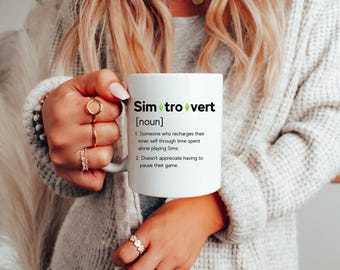 Może przedstawiać: Biały ceramiczny kubek z czarnym napisem Sim-tro-vert [noun] i zielonymi ikonami rombów. Definicja brzmi: 1. Ktoś, kto ładuje swoje wewnętrzne baterie, spędzając czas samotnie na graniu w Sims. 2. Nie lubi przerywać gry, aby ją zapauzować.