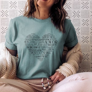 May include: Teal blue t-shirt with a black heart-shaped design featuring a word cloud of essential oils, including "Orange", "Stress Away", "Manuka", "Valor", "Bergamot", "Peace & Calming", "Citrus Fresh", and "Angelica".