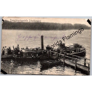 Könnte beinhalten: Vintage Schwarz-Weiß-Postkarte mit einem Dampfschiff, das an der Dampferanlegestelle Schalkendorf angedockt hat. Menschen versammeln sich auf dem Boot und dem Pier. Die Postkarte hat handschriftlichen Text und ist vor einem See- und Baumlinienhintergrund.