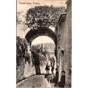 Puede incluir: Postal vintage en blanco y negro con la puerta norte de Totnes. La imagen muestra una puerta de arco de piedra, una escena callejera con gente y el texto "North Gate, Totnes".