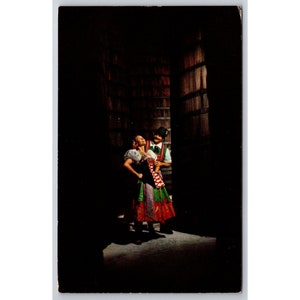 May include: A man and woman in traditional folk costumes stand in a dimly lit room. The woman is wearing a red, green, and white dress with a black bodice. The man is wearing a green vest and brown pants. They are both smiling and looking at each other.
