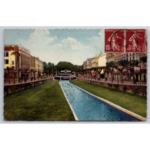 Puede incluir: Una postal antigua que representa un canal que atraviesa una ciudad. El canal está bordeado de árboles y edificios a ambos lados. La postal tiene dos sellos en la esquina superior derecha, ambos con una mujer sosteniendo una antorcha.