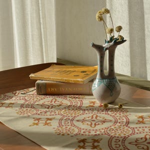 May include: A patterned table runner in cream, red, and orange, with books and a decorative vase. The vase is light pink with blue detailing and holds dried flowers. The books are stacked on the runner, creating a still life.
