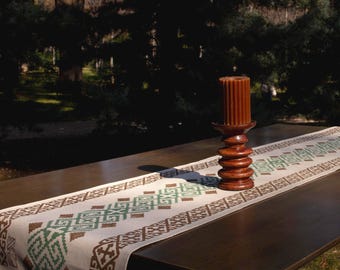 May include: A white table runner with a brown and green geometric pattern. The runner is on a dark brown wooden table with a wooden candle holder and a brown candle on top.