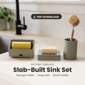 May include: A sage green speckled ceramic sink set including a rectangular sponge holder with a yellow sponge, a soap dish with a white bar of soap, and a cylindrical brush holder containing a wooden dish brush with light brown bristles.