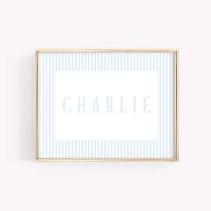 May include: A gold framed print with a light blue and white striped background. The name "CHARLIE" is printed in white on a white rectangle in the centre of the print.