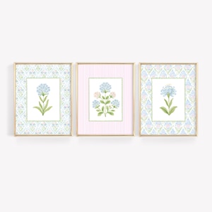 Pode incluir: Três impressões de arte floral emolduradas. Cada impressão apresenta uma ilustração em aquarela de uma flor de hortênsia. As molduras têm bordas douradas. Duas impressões têm um fundo padronizado e uma tem um fundo listrado rosa.