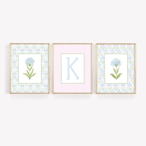 May include: Three framed wall art prints with gold borders. The two outer prints feature a blue flower illustration on a light blue patterned background. The center print displays a large light blue letter K on a pink and white striped background.