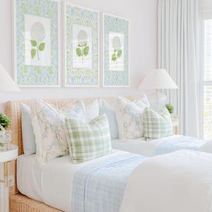 May include: Two twin beds with woven rattan frames, white bedding, and light blue plaid blankets. Decorative pillows feature floral patterns and green gingham checks. Three framed botanical prints of white hydrangeas hang above.