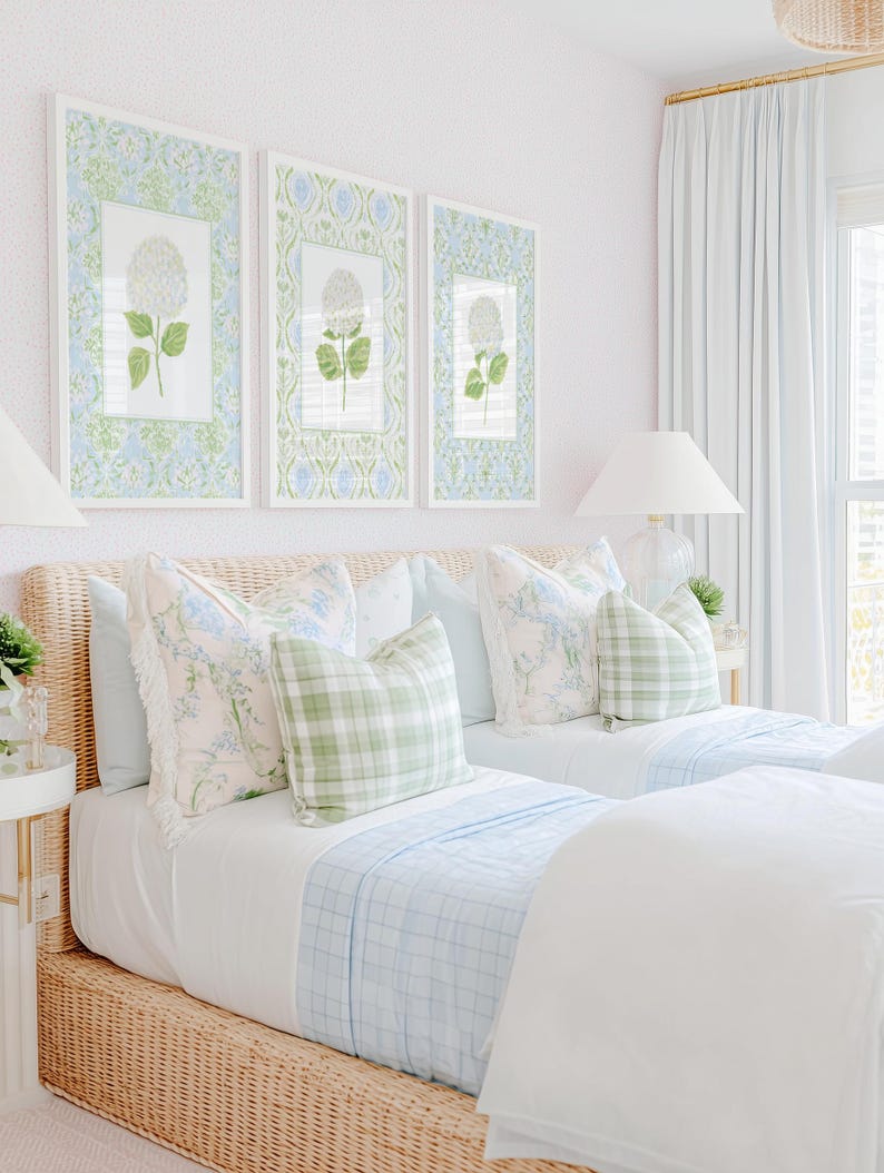 May include: Two twin beds with woven rattan frames, white bedding, and light blue plaid blankets. Decorative pillows feature floral patterns and green gingham checks. Three framed botanical prints of white hydrangeas hang above.