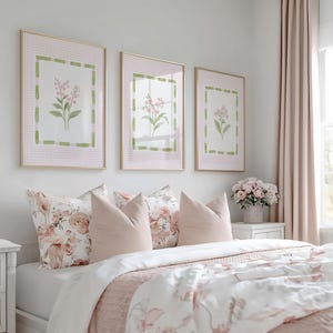 May include: A bedroom scene featuring three framed floral prints above a bed with pink and white bedding. The bed has several pillows in shades of pink and floral patterns. A white nightstand with a lamp is on the left.