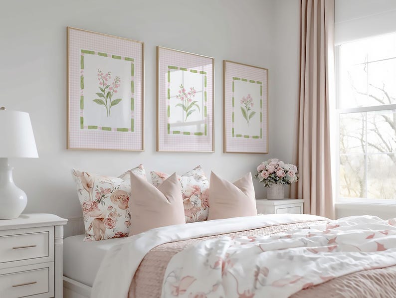 May include: A bedroom scene featuring three framed floral prints above a bed with pink and white bedding. The bed has several pillows in shades of pink and floral patterns. A white nightstand with a lamp is on the left.